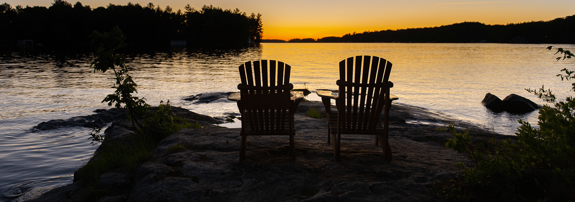 Adirondack chairs on a rock facing a lake at sunset