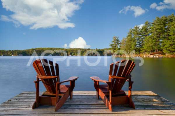 Two Adirondack chairs on a wooden dock facing a lake in Muskoka - GettaPix